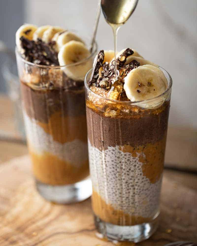 45 degree shot of 2 glass jars with chia pudding, peanut butter and chocolate topped with fresh banana and honey being drizzled on