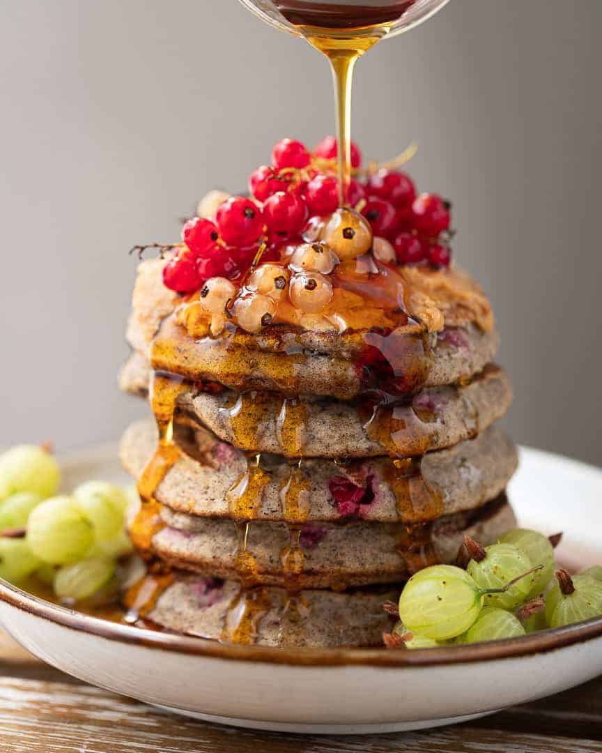 stack of gluten free pancakes topped with currants and maple syrup being poured over