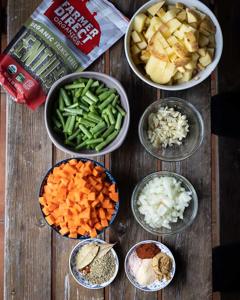 Birds eye view of a wooden table with bowls of diced carrots, onions, garlic, potatoes, lentils, green beans and spices