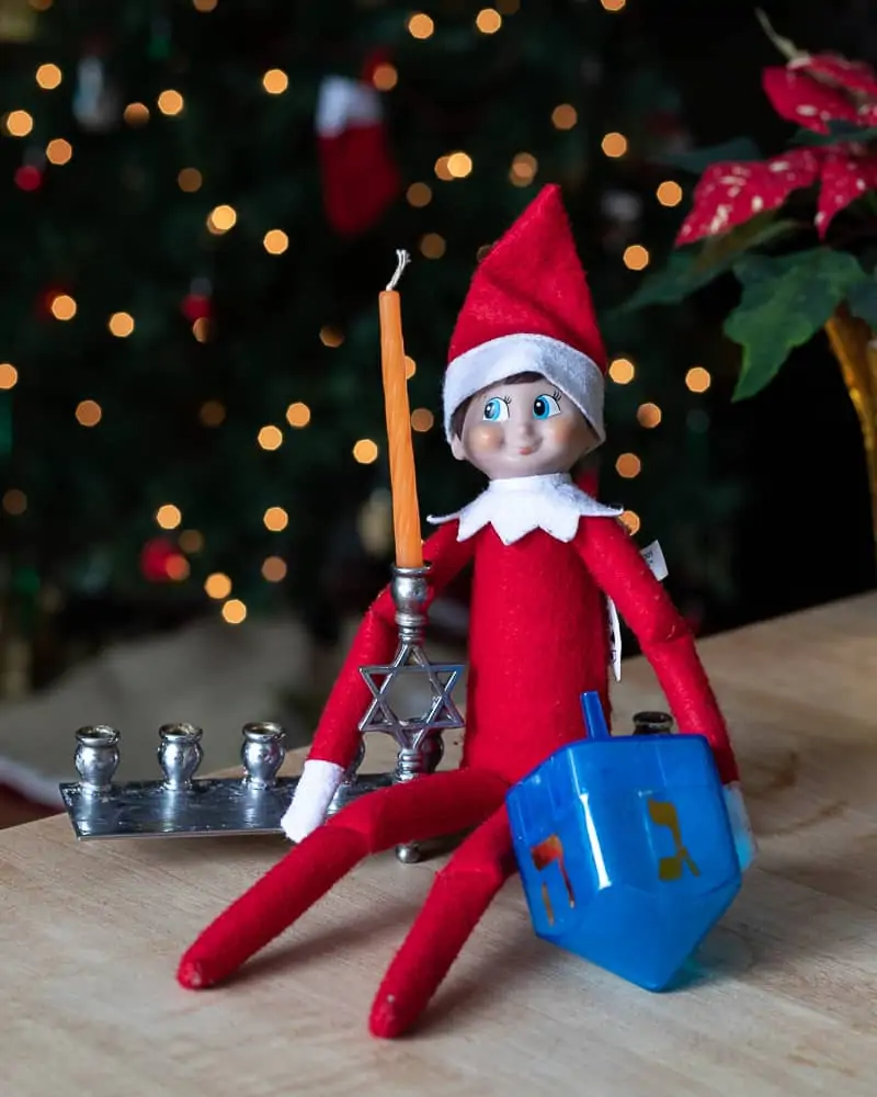 Elf on a shelf on a menorah holding a dreidel for a post on interfaith families