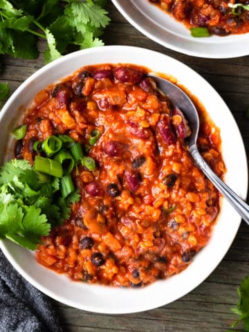 Image of a delicious bowl of bean chili on a white plate, garnished with fresh cilantro, sitting on a wooden table. The chili is rich and hearty, with chunks of onions, tomatoes and beans in a thick sauce.