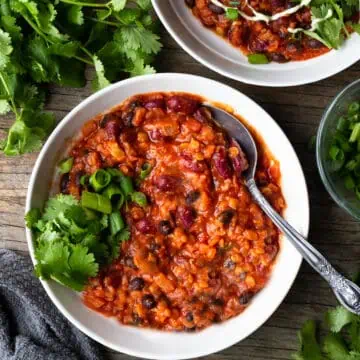 Image of a delicious bowl of bean chili on a white plate garnished with fresh cilantro. The bowl is sitting on a wooden table and is for a blog post on a recipe for simple chili. The chili is rich and hearty, with chunks of onions, tomatoes, and beans in a thick sauce.