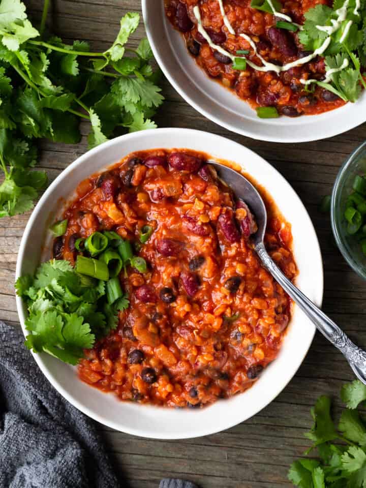 Image of a delicious bowl of bean chili on a white plate garnished with fresh cilantro. The bowl is sitting on a wooden table and is for a blog post on a recipe for simple chili. The chili is rich and hearty, with chunks of onions, tomatoes, and beans in a thick sauce.