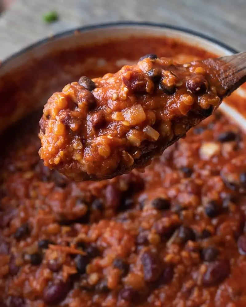 Image of a delicious pot of chili sitting on a wooden table. There is a closeup of a wooden spoon full of beans and sauce. 