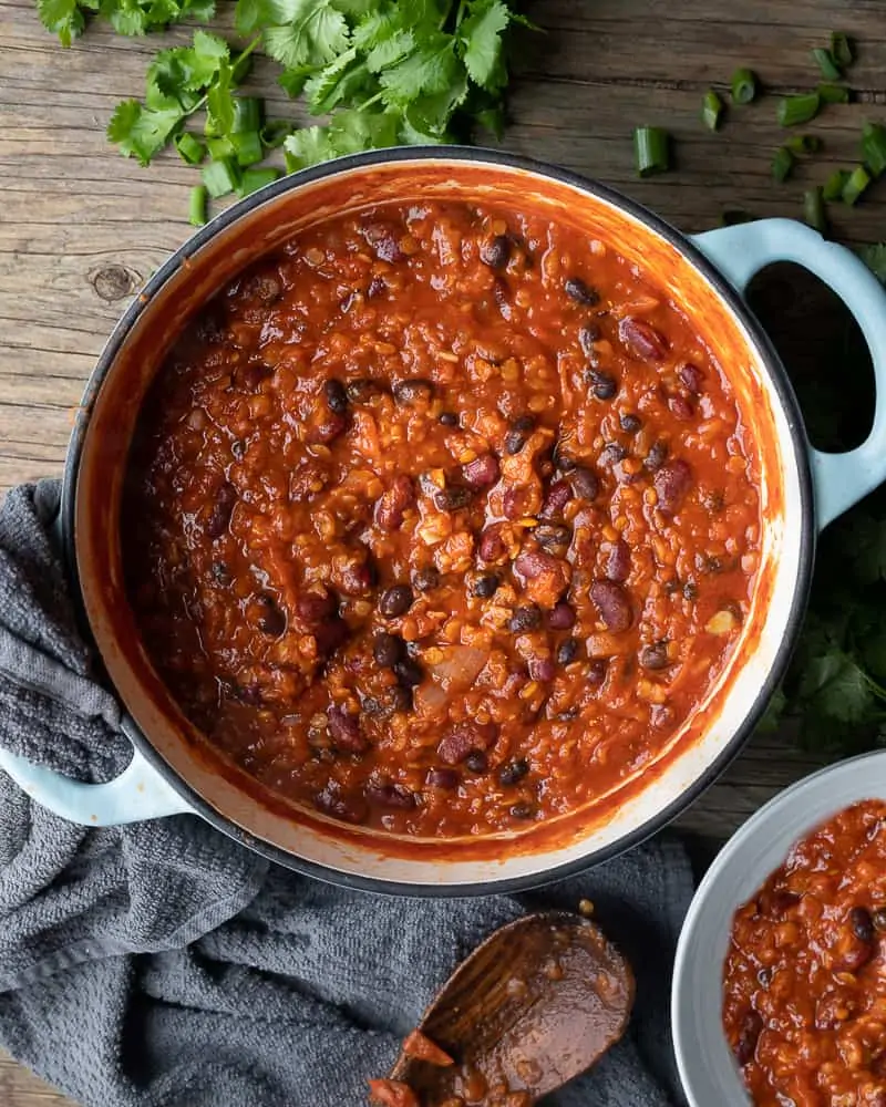Overview image of a simple recipe for chili, the image has a pot of bean chili sitting on a wooden table. The pot is surrounded by a wooden spoon, a grey kitchen towel, and fresh cilantro. The chili is rich and hearty, with chunks of onions, tomatoes, and beans in a thick sauce.