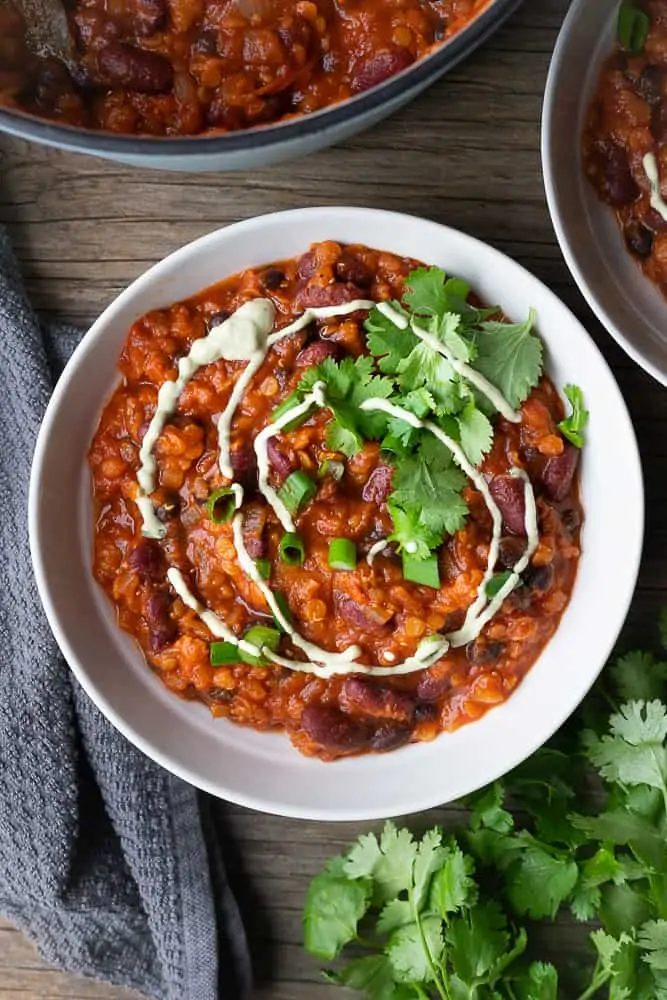 Image of a delicious bowl of easy stovetop chili on a white plate garnished with fresh cilantro and a creamy sauce. The bowl is sitting on a wooden table. The chili is rich and hearty, with chunks of onions, tomatoes, and beans in a thick sauce.