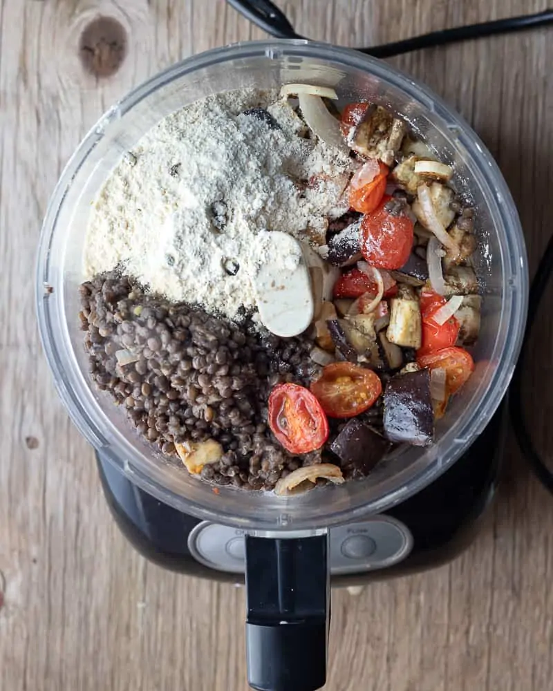 Close-up view of a food processor with lentils, chickpea flour, roasted tomato, onions, and eggplant for a recipe on meatless meatballs.