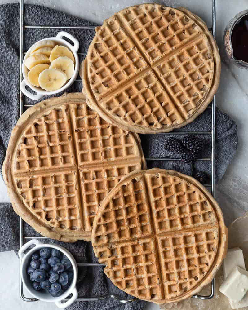 3 waffles cooling on a rack surrounded by banana, blueberries and maple syrup