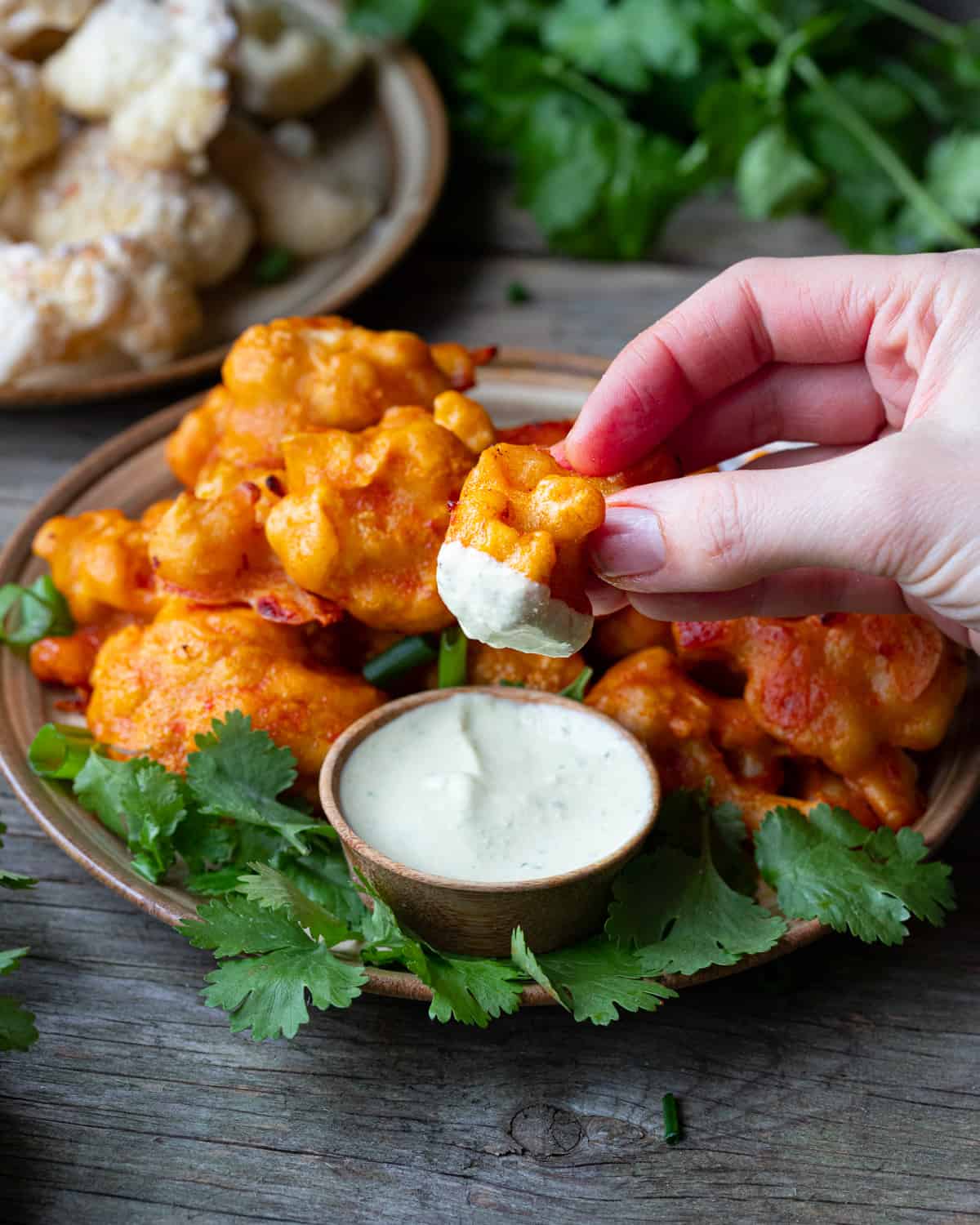 Image of a bowl of vegan wings in a vegan buffallo sauce. There is a human hand dipping a wing into a vegan creamy dressing to show that this recipe is good with sauces.