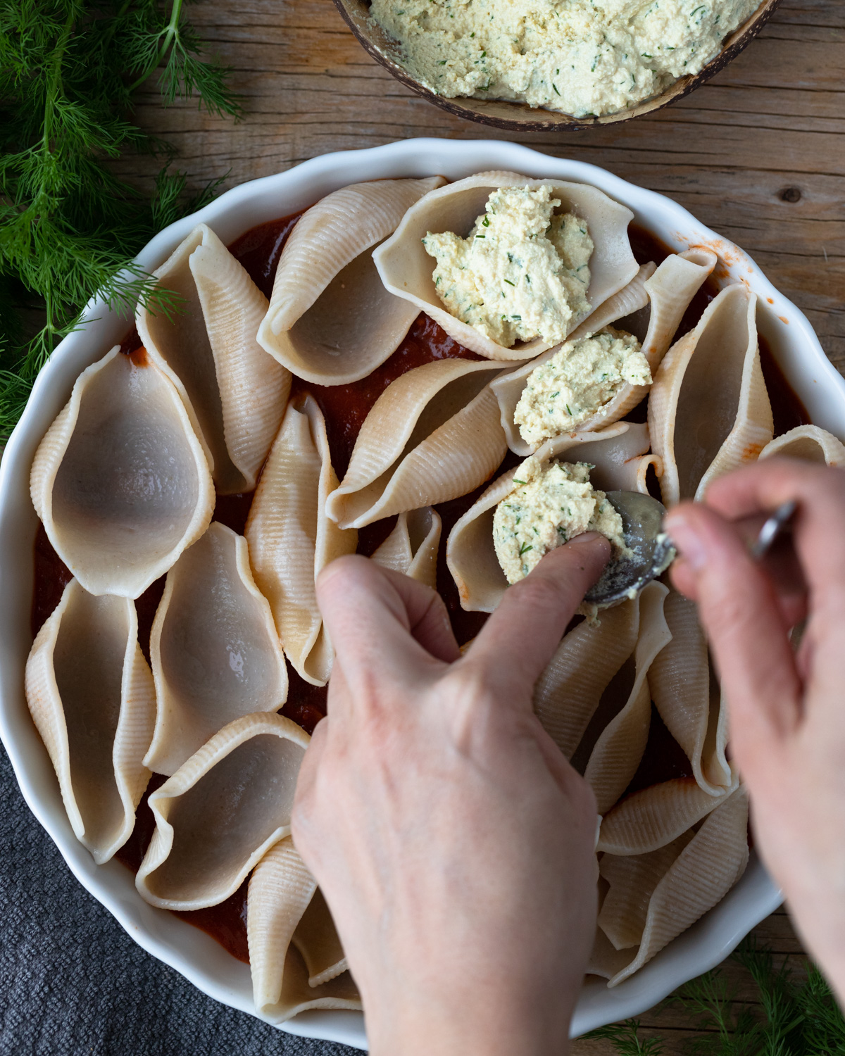 Top-down view of a plate of unbaked gluten-free pasta shells being stuffed with vegan ricotta. The dish is on a wooden table with fresh dill and a napkin.