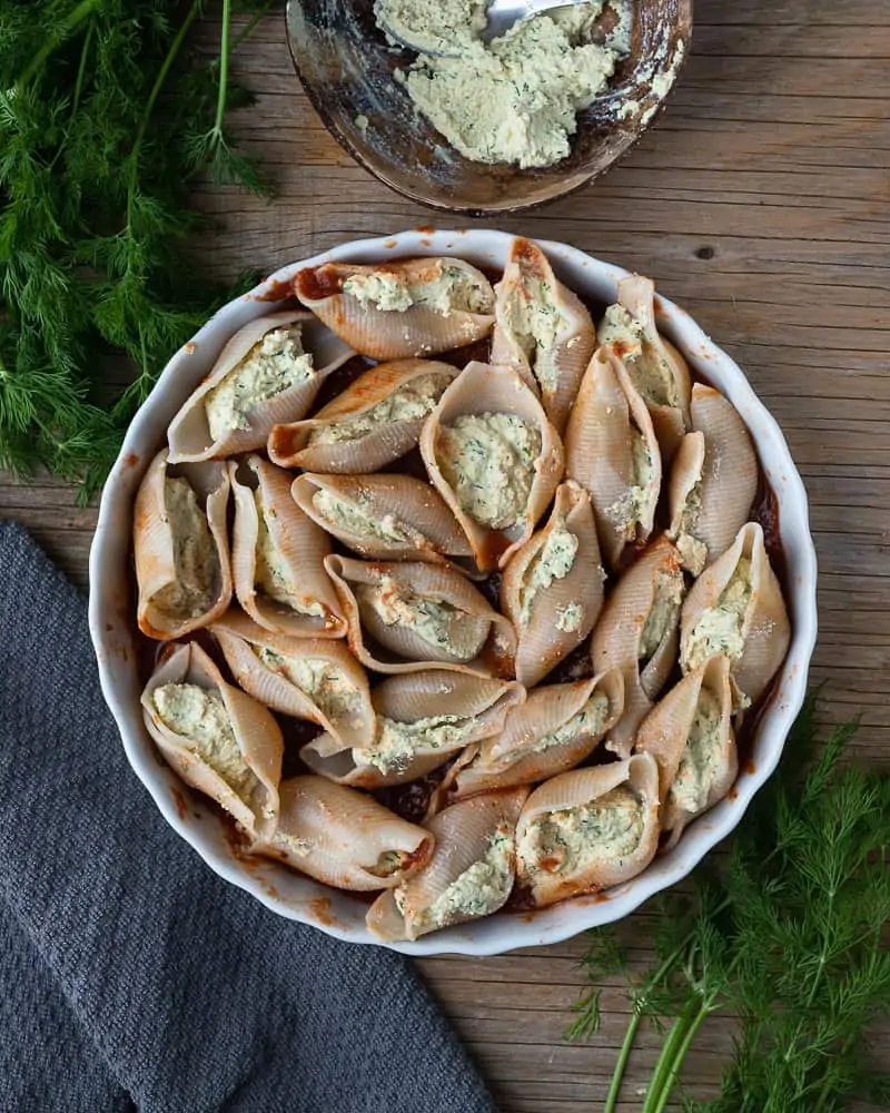 Top-down view of a plate of unbaked gluten-free pasta shells stuffed with tofu ricotta ready for the sauce. The dish is on a wooden table with fresh dill and a napkin.