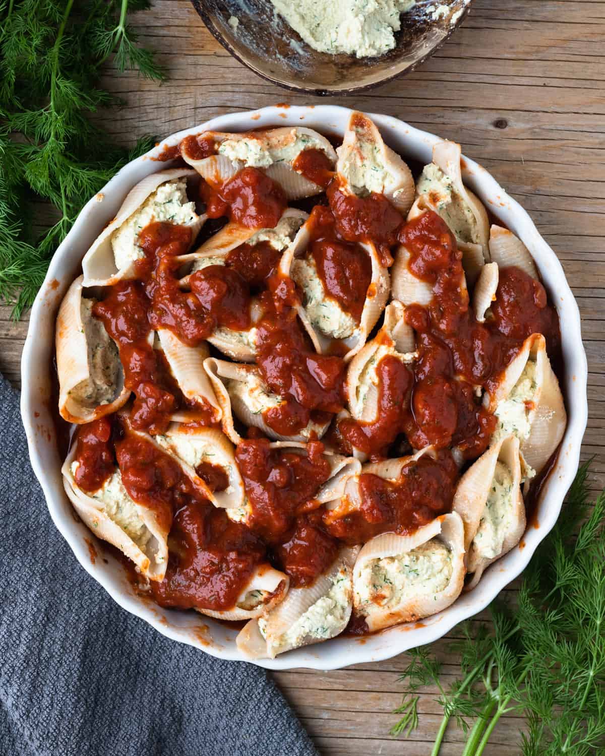 Top-down view of a plate of unbaked gluten-free pasta shells stuffed with tofu ricotta in tomato sauce. The dish is on a wooden table with fresh dill and a napkin.