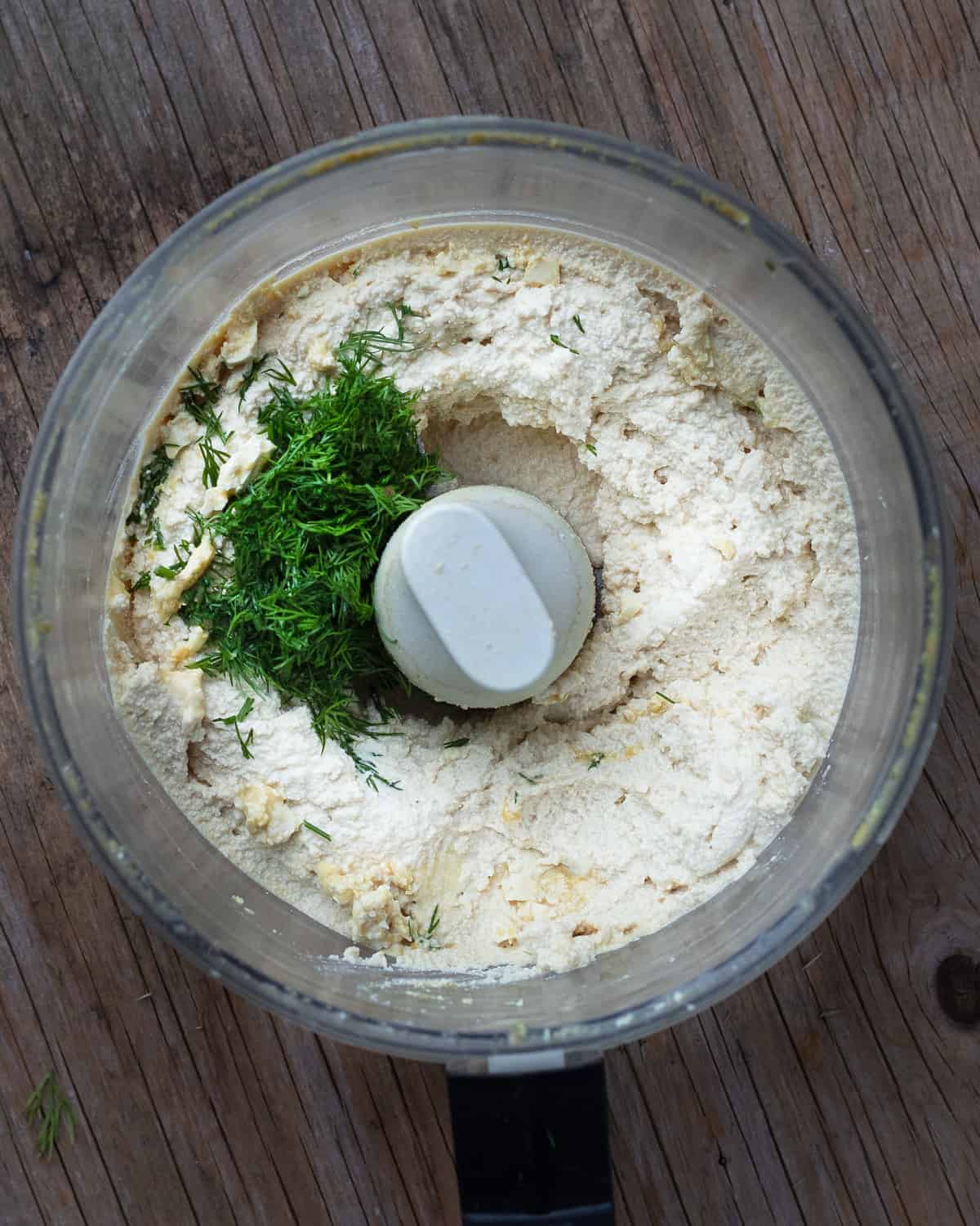 Overhead shot of a food processor on a wooden table with tofu, spices and, and parsley inside that has been processed into a vegan cheese