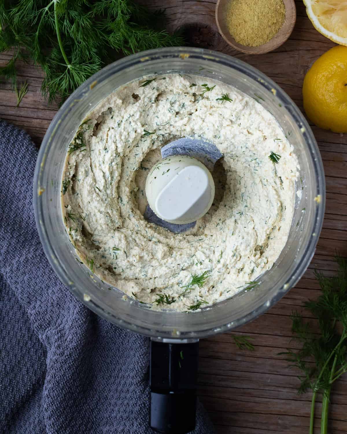 Overhead shot of a food processor on a wooden table with tofu, spices, and, and parsley inside that has been processed into a creamy vegan ricotta cheese