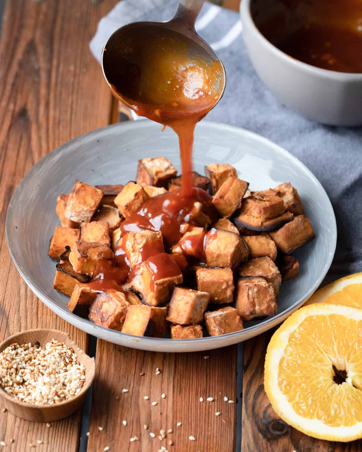 Close-up of a bowl of roasted and cubed tofu. A metal spoon is pouring orange sesame sauce on top of the tofu. The bowl is sitting on top of a wooden table