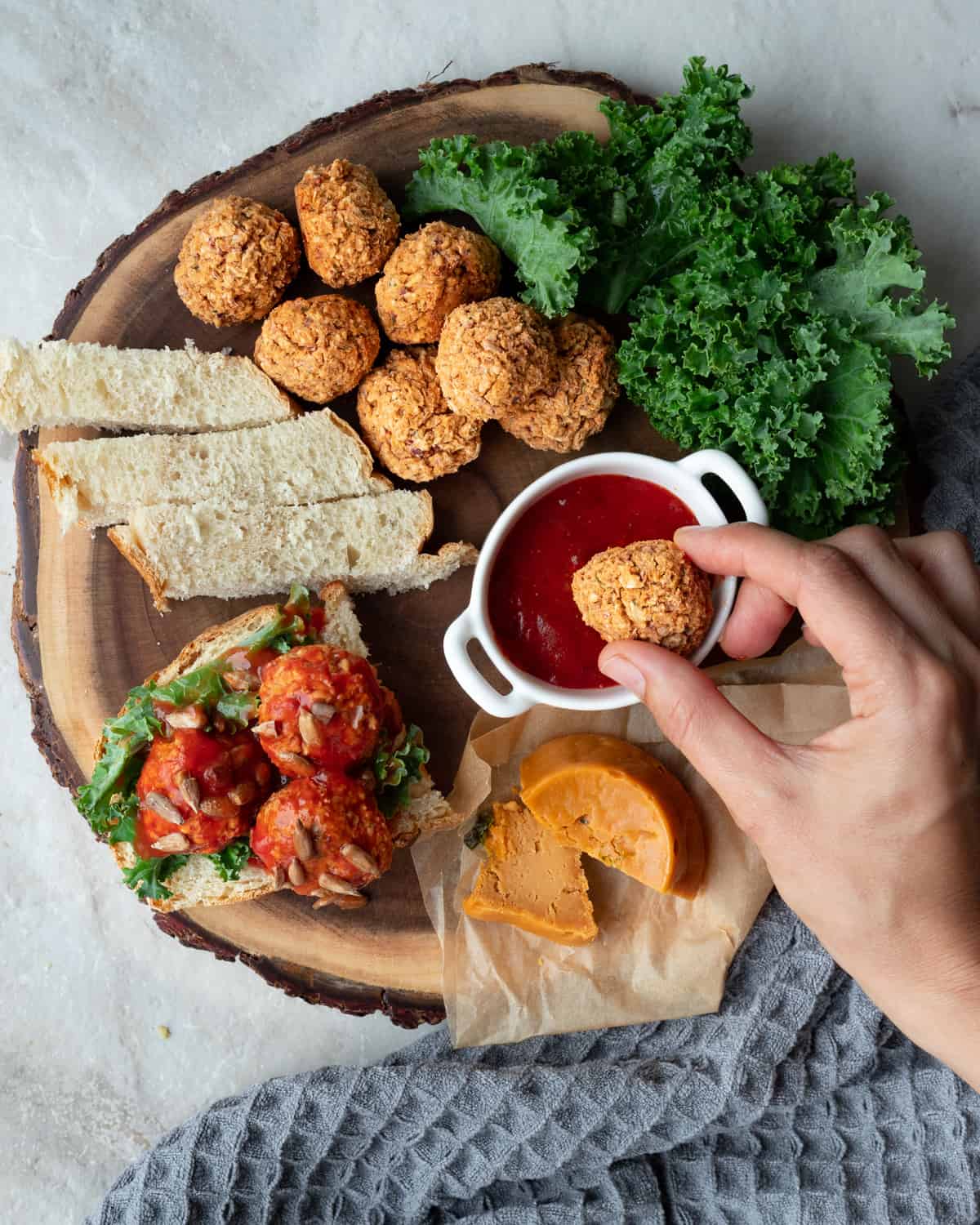 Top-down view of a cutting board full of vegan cheese, kale, toast, and vegan meatballs. There is a human hand dipping a meatball into a small bowl of tomato sauce at the center of the cutting board. There is a grey napkin at the bottom of the photo.