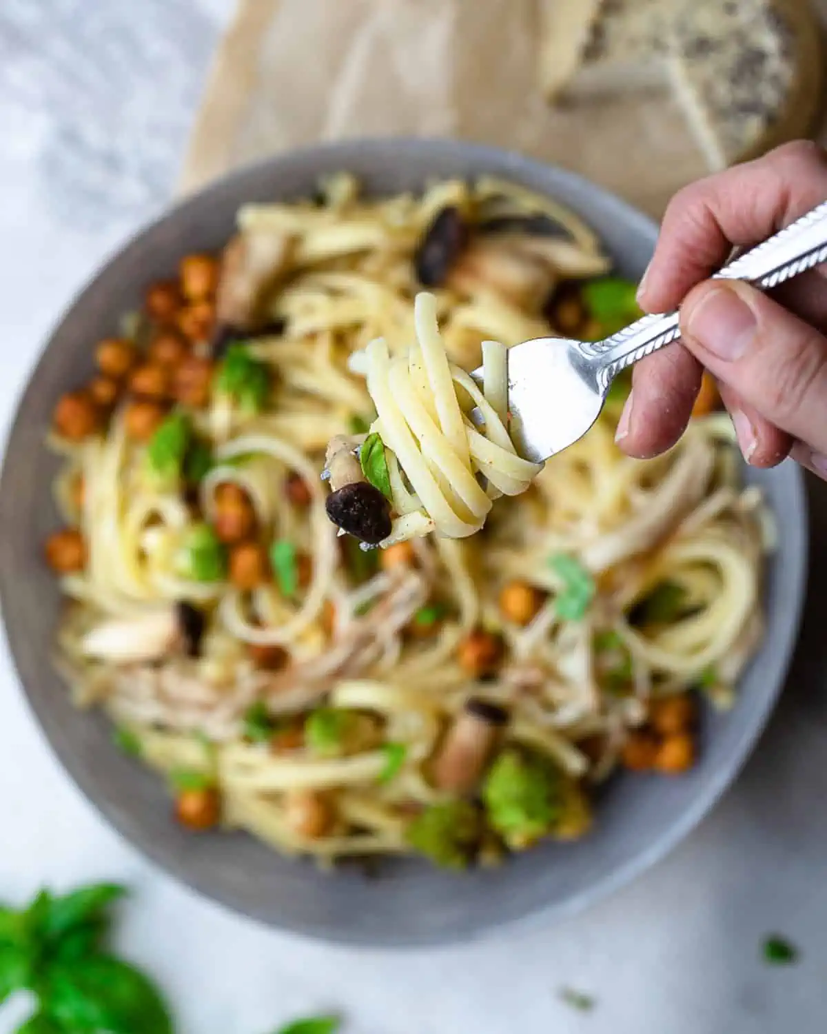 Close up of a fork twirled with gluten free fettucini pasta. There is a bowl out of focus in the background with roasted mushrooms, chickpeas, and romanesco cauliflower topped with a creamy sauce.
