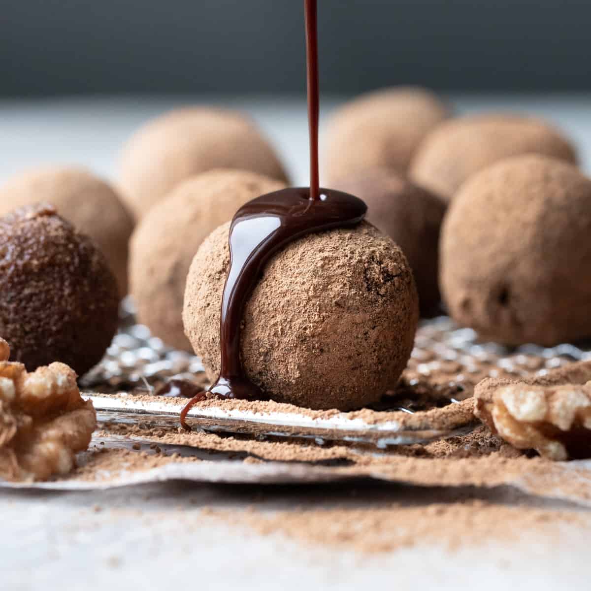 Image of a energy bites sitting on a wire rack being drizzled with chocolate syrup. There are other bites sitting out of focus behind. Image is for how to make date balls.