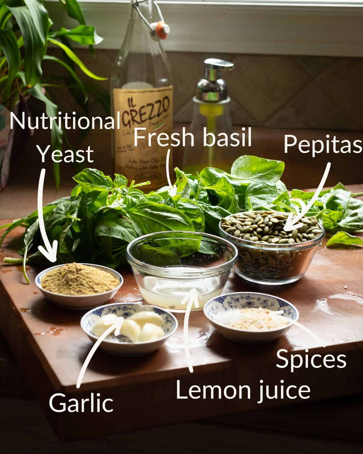 Ingredient shot for a post on how to make nut-free pesto sauce. The image has a wooden cutting board with small bowls of garlic, spices, nutritional yeast, lemon juice, and pepitas.