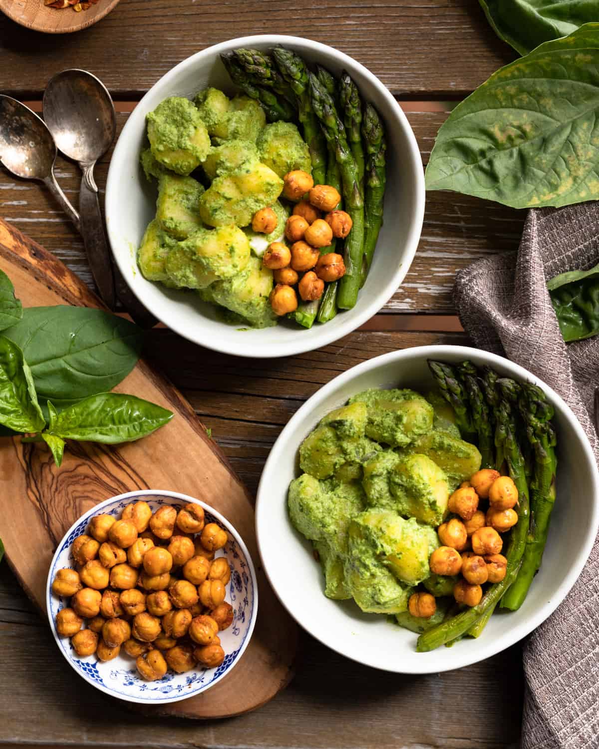 Top down view of a table with bowls of gnocchi covered in a pesto without pine nuts sauce. The pasta is topped with roasted chickpeas and aspargus. There is basil around and a small bowl of the chickpeas on the table as well.