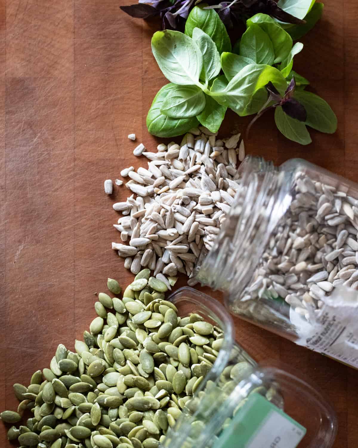 A wodden cutting board with 2 glass jars. The jars are full of pumpkin seeds and pepitas to show the difference between the type types of seeds.