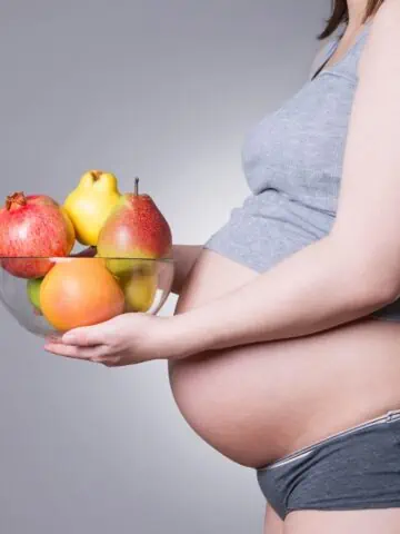 Expecting mother cradling her belly while holding a vibrant plate of colorful plant-based foods. Image if for a blog post on the safety of vegan and vegetarian diet plans