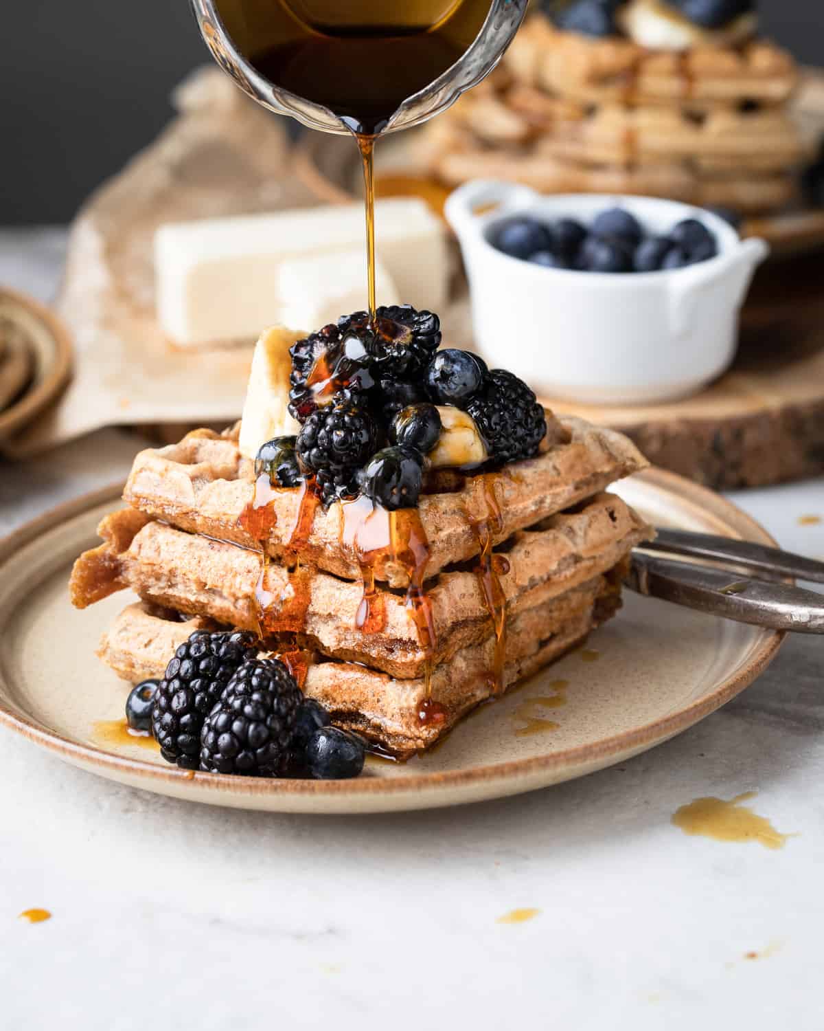 A stack of dairy free waffles with maple syrup being drizzled on top. The waffles have fresh fruit on top and is for a recipe post.