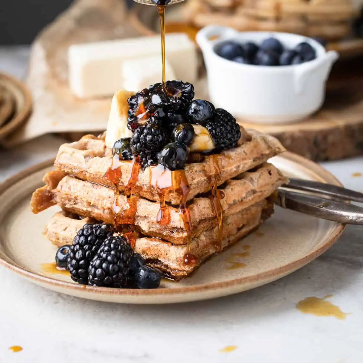 A stack of waffles with maple syrup being drizzled on top. The waffles have fresh fruit on top and is for a recipe post.