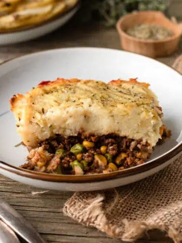 Image for a recipe post on how to make plant-based shepherd's pie. The image shows full plates of the pie being on a wooden table.