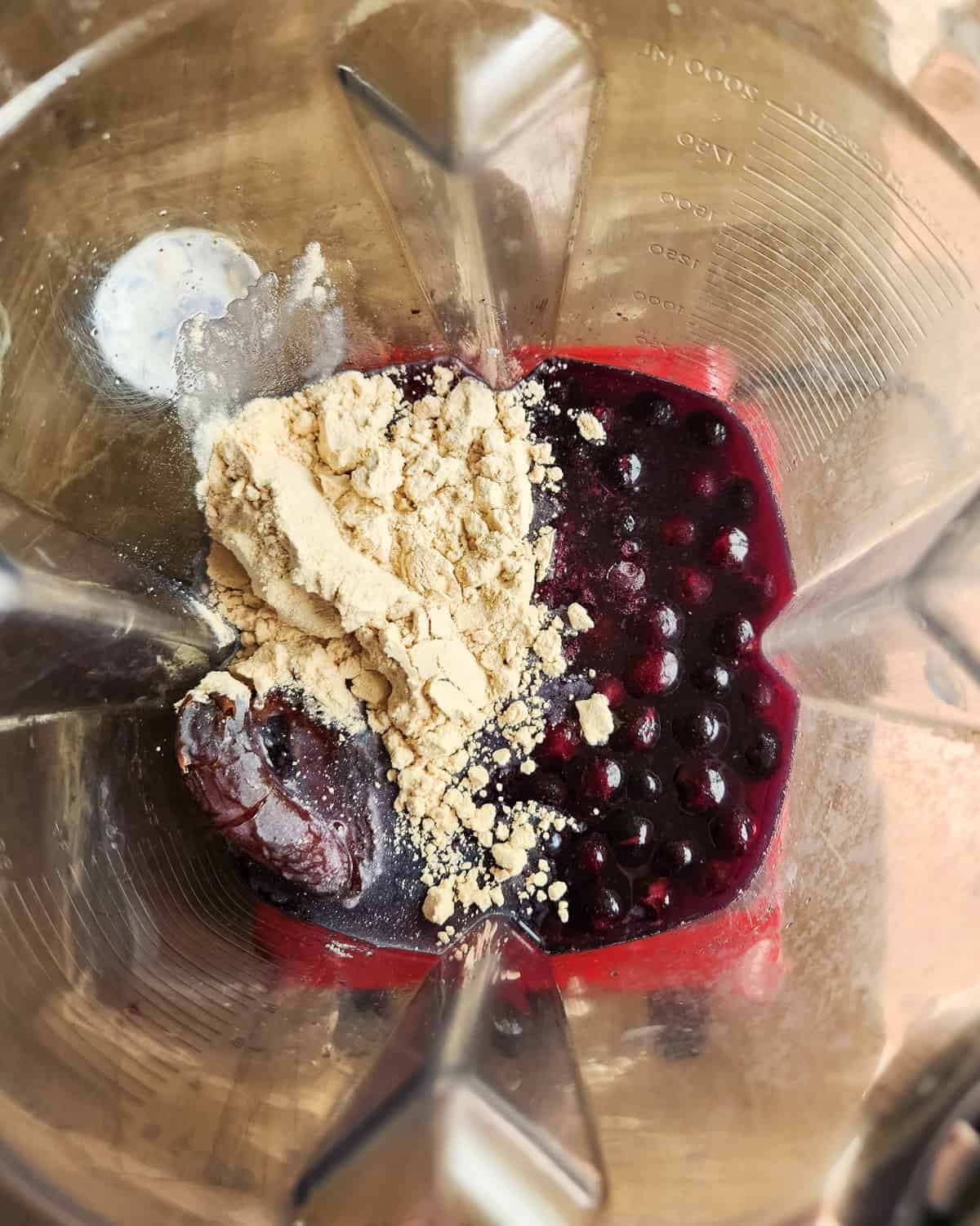 Image shows the inside of a high speed blender with frozen blueberries, protein powder, dates and water. The image is a process image to show how to make a high protein smoothie.