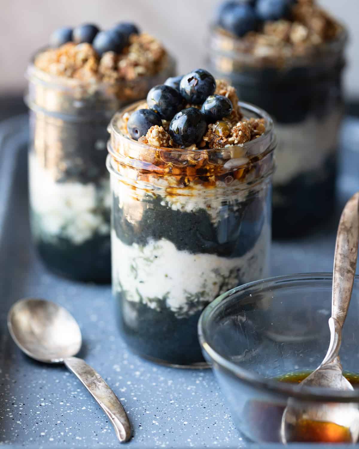 Close up image of a mason jars with layers of a high protein smoothie and overnight oats topped with fresh granola and blueberries. There are 2 more high protein overnight at parfaits in the background and a bowl of maple syrup in the foreground.