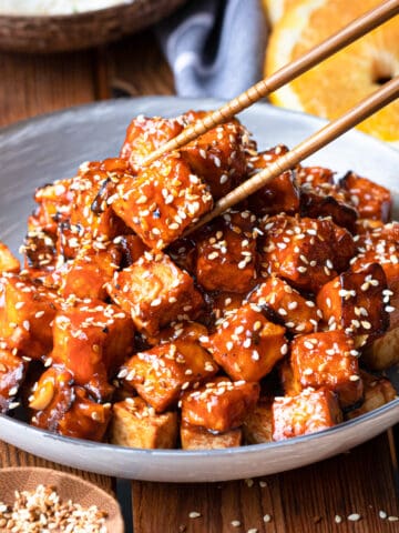 A cozy image of a bowl of orange tofu. the orange is sticky and cubed and sprinkled with roasted sesame seeds.