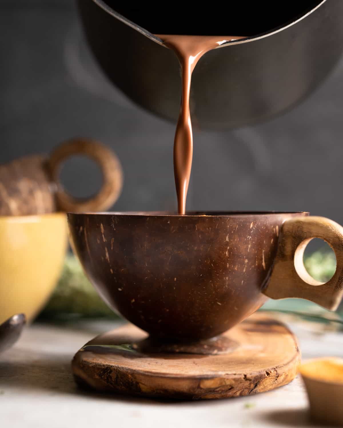 Image of hot chocolate being poured into a wooden mug that is sittig on a wooden cutting board.