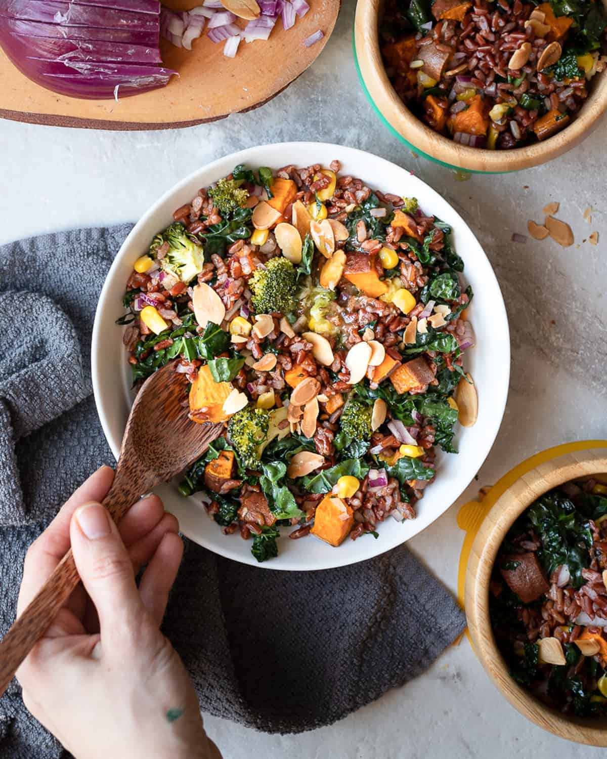 Top down image image of a bowl of roasted veg and red rice salad topped in a gluten free salad dressing. The image shows a human hand with a spoon mixing the salad.