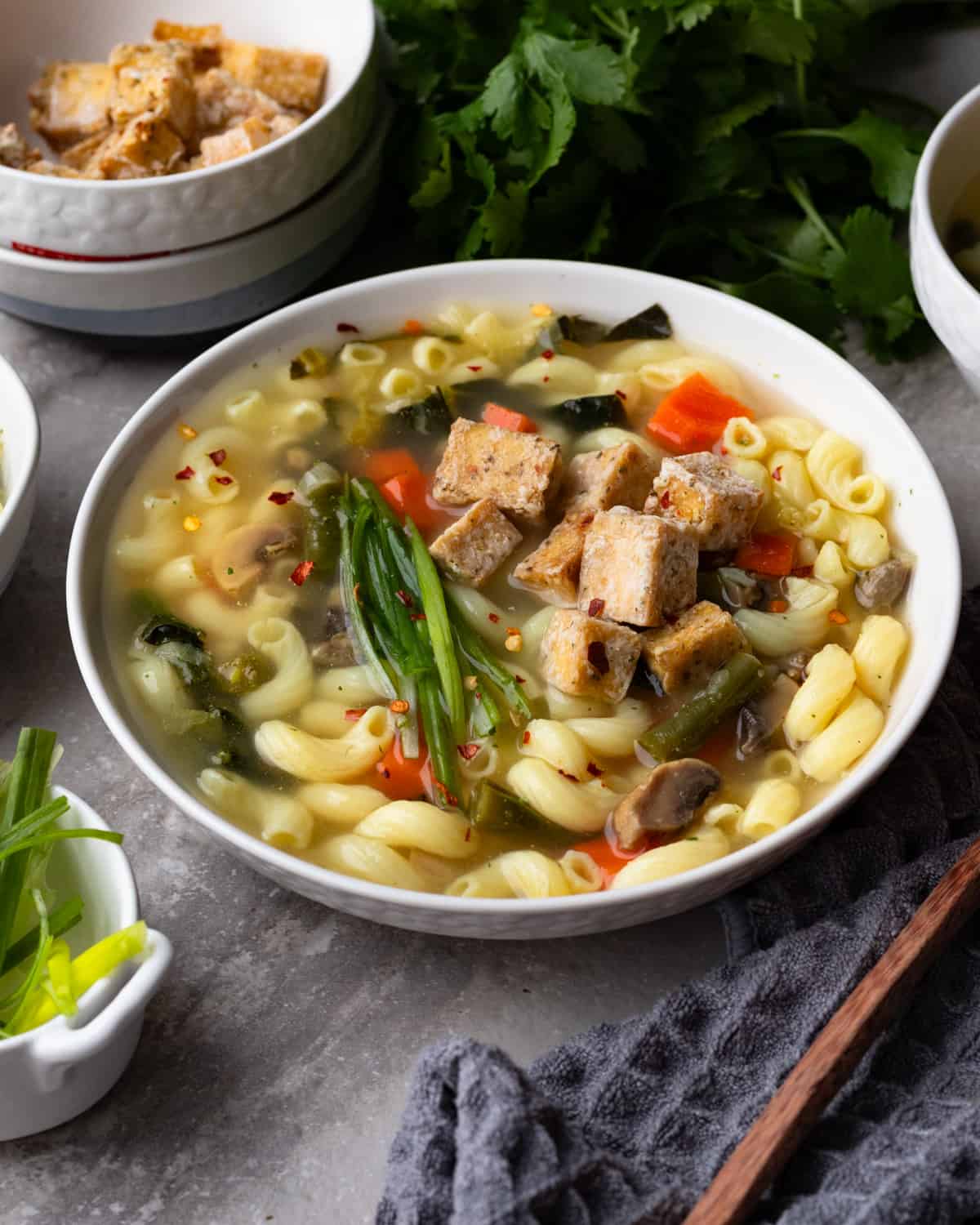 Cozy image of a bowl of vegan noodle soup made with a vegetable broth with gluten free noddles, tofu, chickpeas, carrots and mushrooms. The smaller bowls have green onions and another soup bowl.