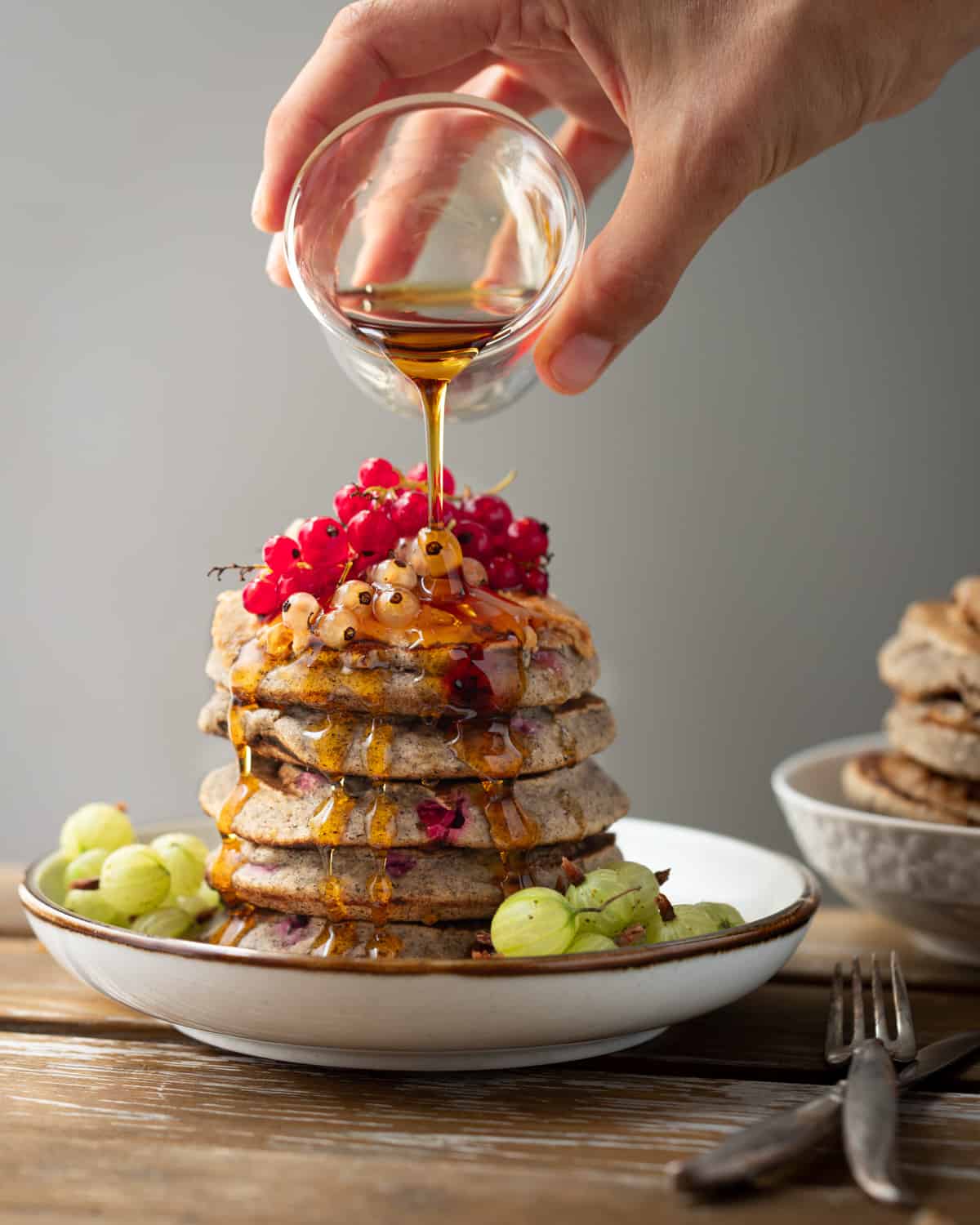 Image of a stack of pancakes topped with fresh currents. There is maple syrup being poured on top of the stack. 