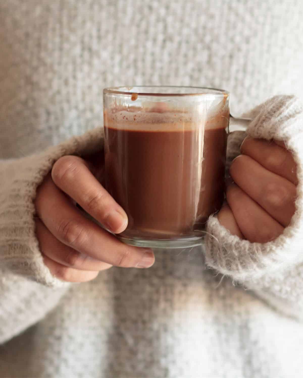 Image of a human holding a mug of protein hot chocolate made without milk. The women holding the mug is wearing a white cozy sweater.