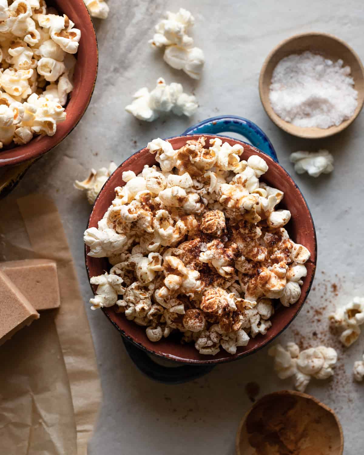 Image of a bowl of popped cinnamon vegan popcorn. The bowl is surrounded by some vegan butter, and small bowls of ground cinnamon and sea salt.