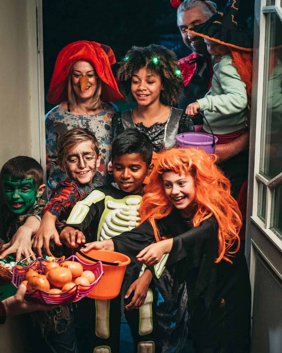 Image of 6 children and 2 adults dressed in Halloween costumes. They are standing at a door trick or treating and are grabbing for a basket of candy and tangerines.