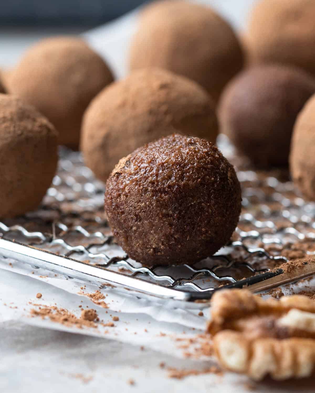 This image features a moist-looking bliss ball with a rich, dark color contrasting against the lighter cocoa-dusted bliss balls in the background. The focus on the darker ball in the forefront, resting on a wire cooling rack, emphasizes its unique texture, indicative of the variety of healthy ingredients used to make these bliss balls appealing to kids.