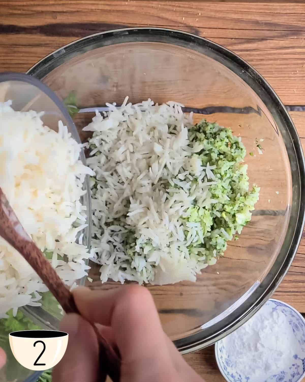 Uma tigela de vidro com mistura de arroz processado e uma mão humana adicionando mais arroz cozido. Esta é a etapa 2 das instruções de como fazer bolinhos de arroz assados.
