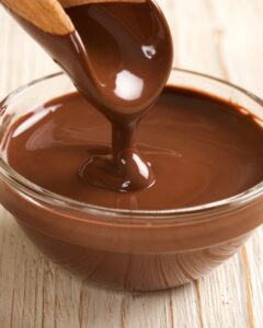 A glass bowl of melted chocolate being stirred with a wooden spoon to create a smooth creamy sauce.