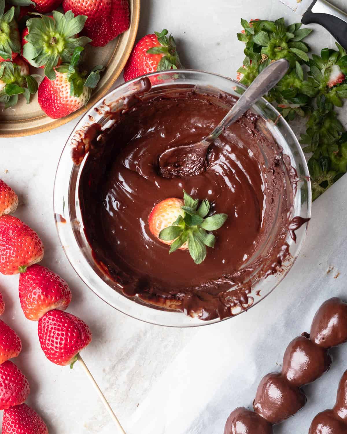 A glass bowl of dark melted chocolate with a strawberry being dipped inside.