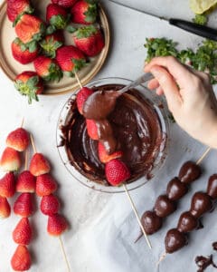 A bowl of melted dark chocolate with a kebob of skewered fresh strawberries over top the bowl about to be drizzled with the chocolate.