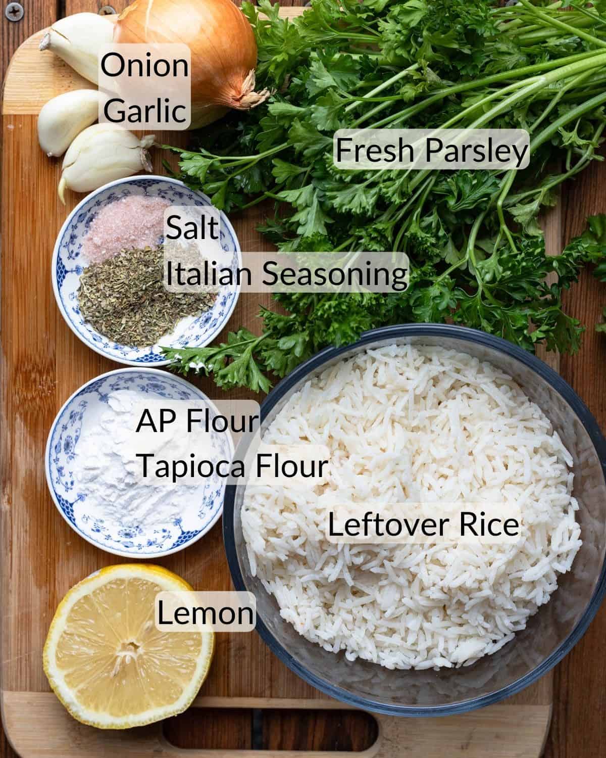Ingredients needed to make baked rice balls vegan. There is a wooden cutting board with fresh lemon, a bowl of leftover rice, tapioca flour, fresh parsley, fresh onion, fresh garlic, flour, salt and Italian seasoning.