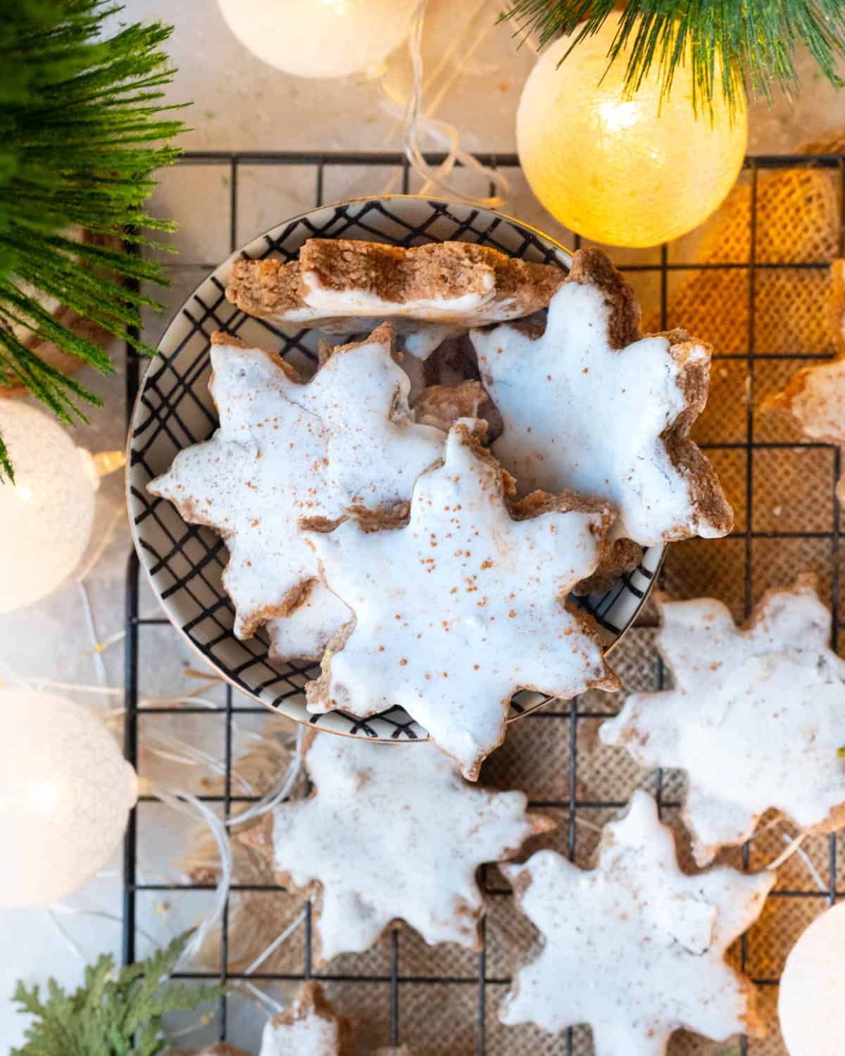 Vegan Zimtsterne cookies with a delicate white icing, resting on a cooling rack with twinkling lights in the background