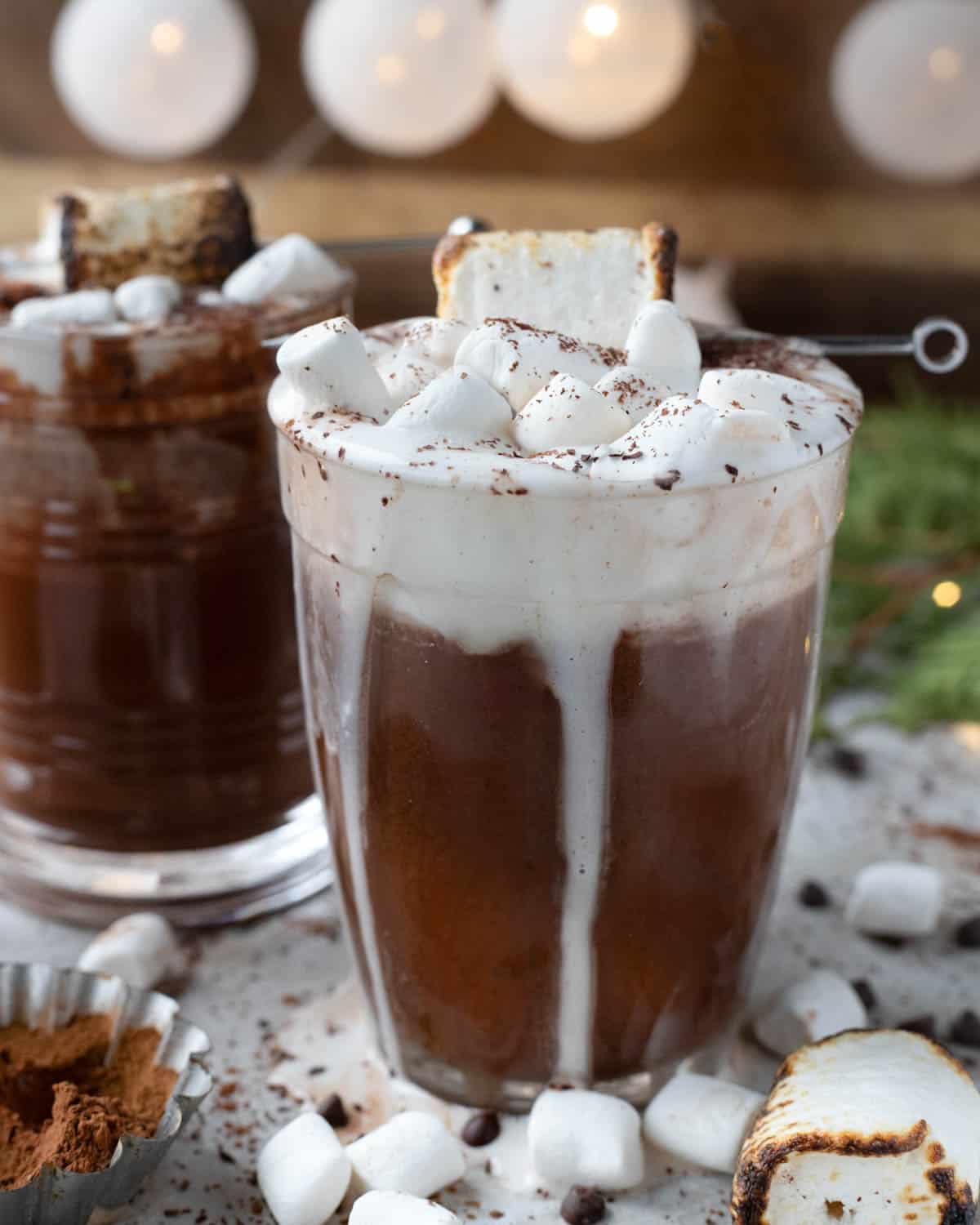 A festive setting featuring a tall glass of hot chocolate with oat milk, crowned with a swirl of whipped cream and marshmallows. The background is softly lit, suggesting a cozy ambiance.