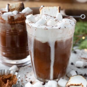 A close up of a glass of oat milk hot chocolate, elegantly presented with a creamy top, chocolate drizzle, and a toasted marshmallow, suggesting a luxurious treat.