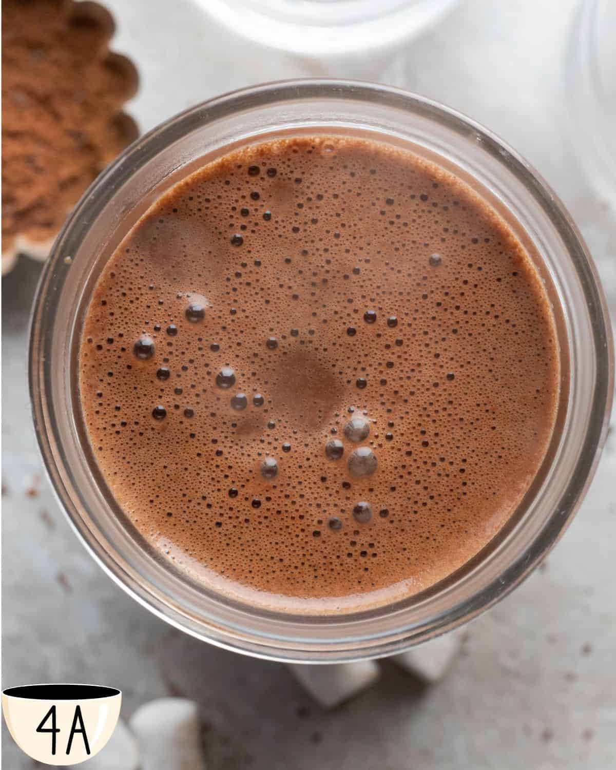 A glass jar viewed from above, filled with rich and smooth oat milk hot chocolate. The surface is covered with small, delicate bubbles from the warmth of the freshly mixed beverage, indicating it's ready to be enjoyed.
