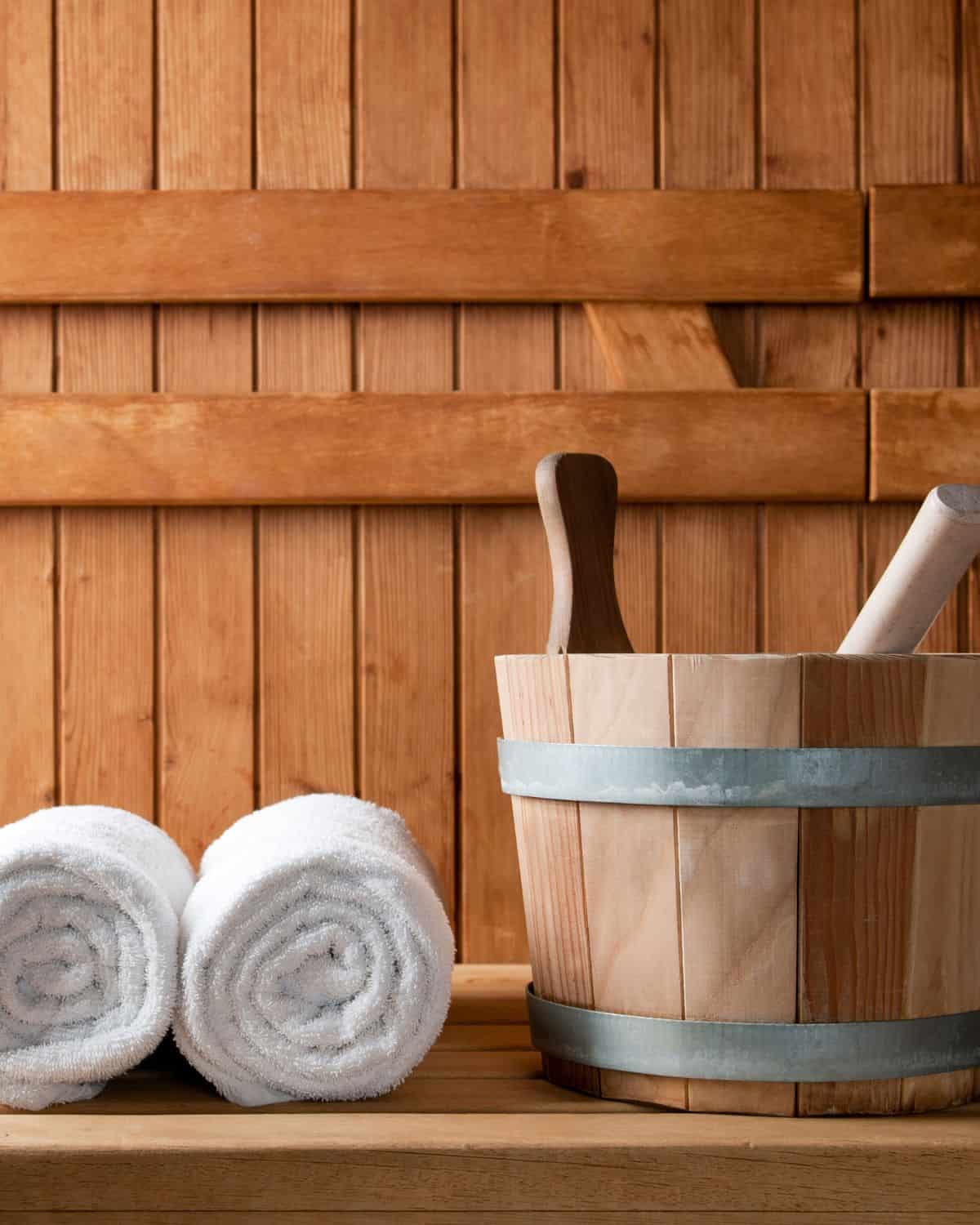 A bucket with a wooden spoon and two towels sitting on a cedar bench inside a traditional sauna.